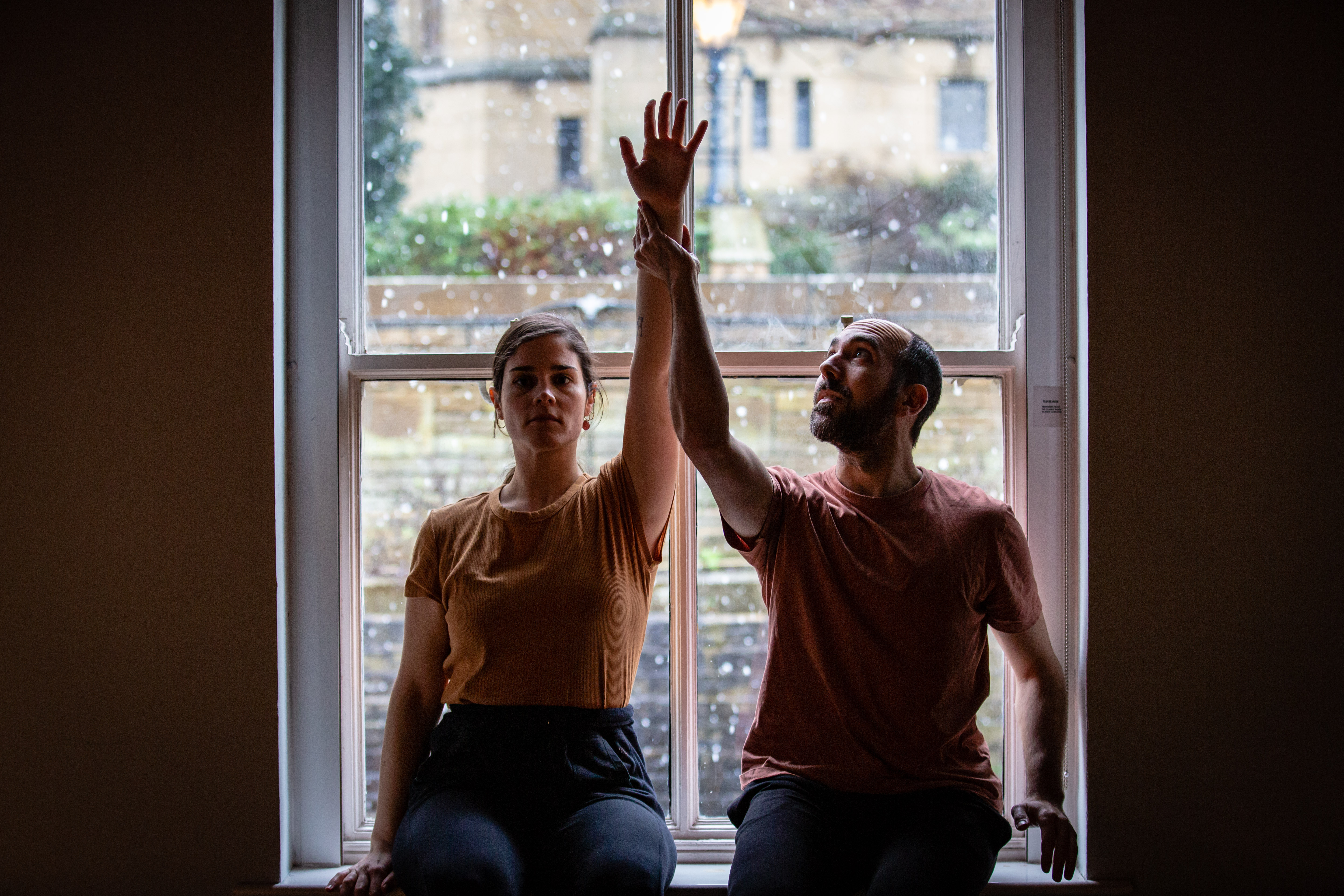 A man and a woman sit side by side in front of a window, each raising a hand above their heads to meet in the middle.