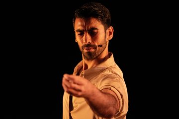 A south asian man, holding his left arm in front of him in front of a black background