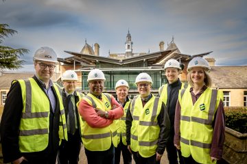 A group of seven people wearing hi-vis jackets and hard hats stand in front of a building