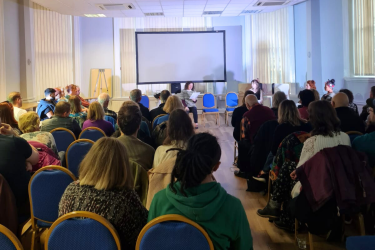 an audience watching a performer who is sat down reading a script