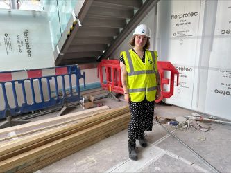 A white woman wearing a hard hat and hi-vis vest stands in on a building site.