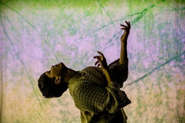 A dancer wearing a wool jumper posing in front of a projection of some trees